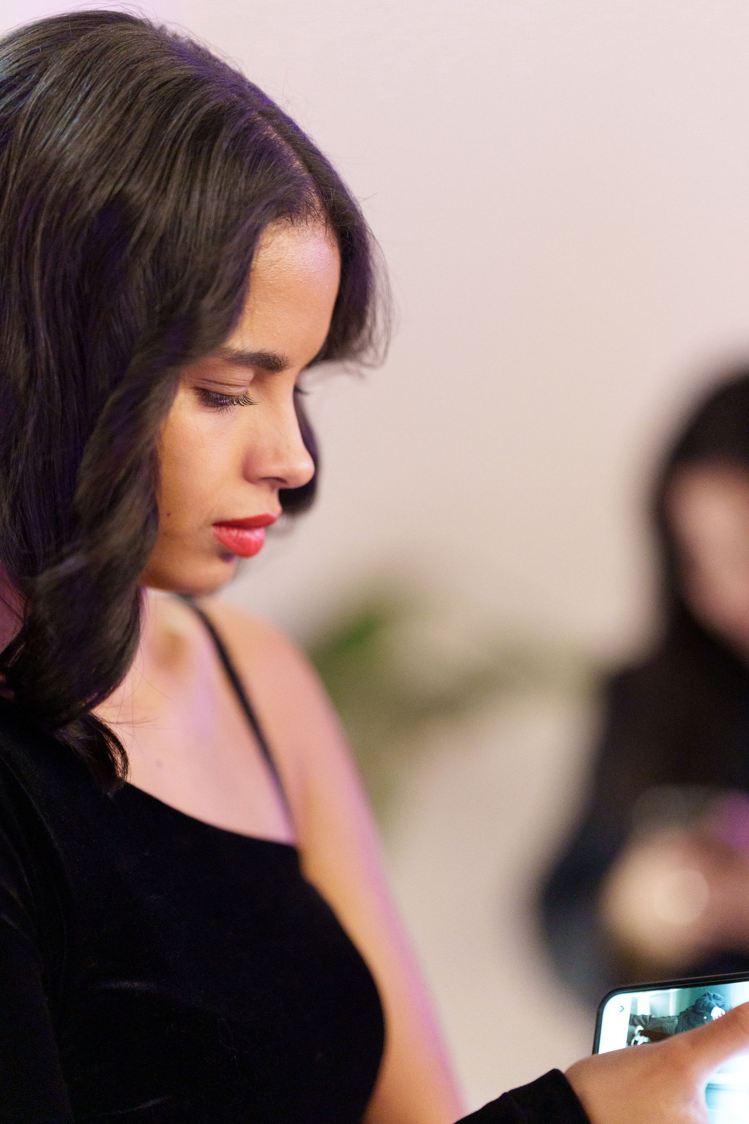 Young woman with dark hair and red lips looks at her smartphone while wearing a black dress at an indoor event.