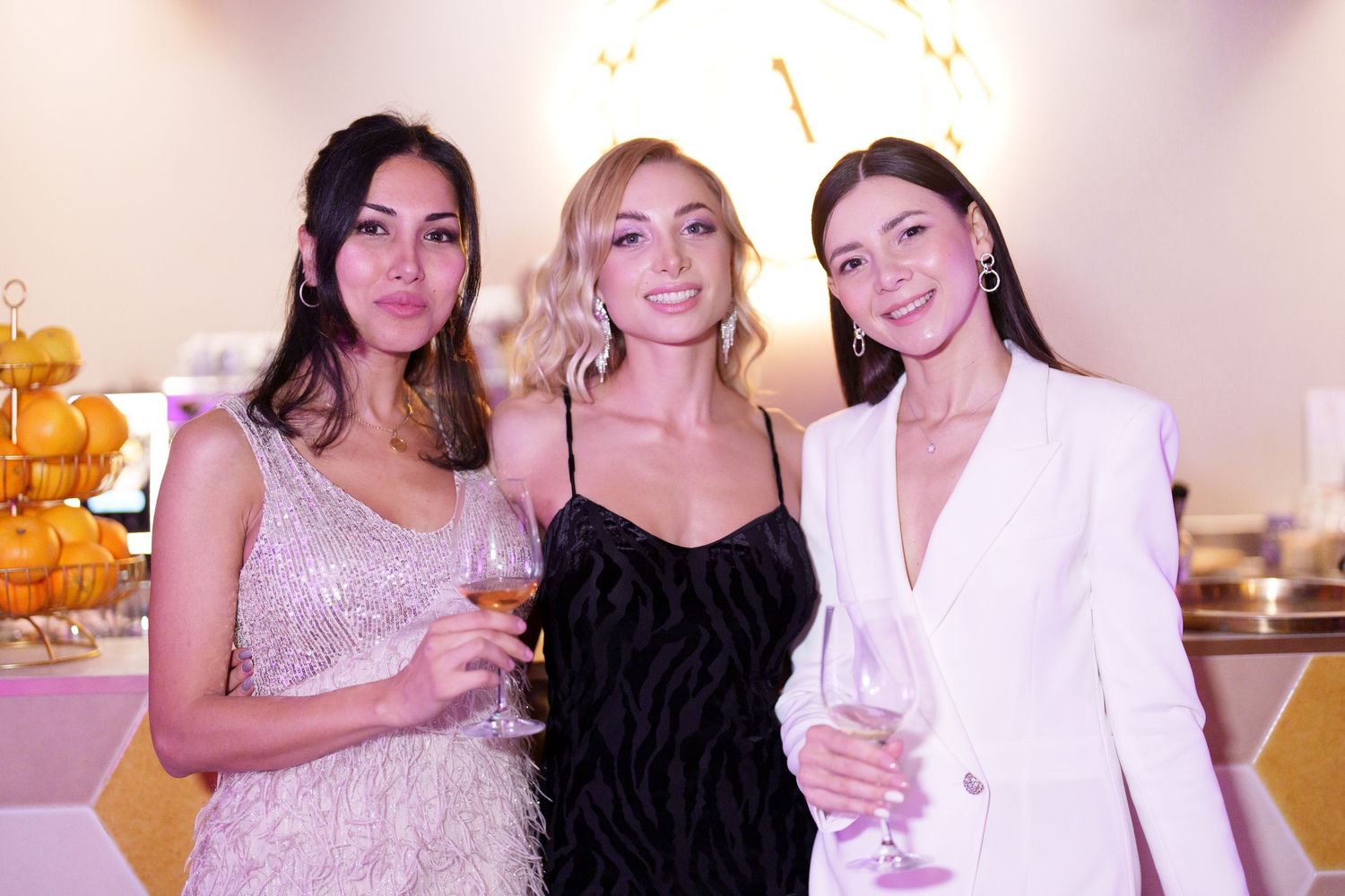 Three elegant women in formal attire smile while holding champagne glasses at a upscale event.