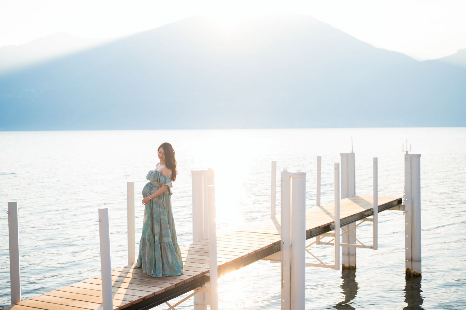 Pregnant woman in teal gown stands on wooden dock overlooking calm lake with mountain backdrop in soft sunlight.