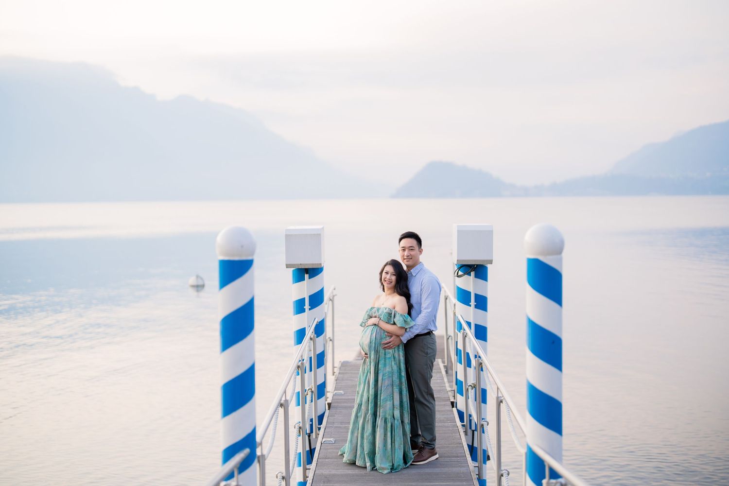 Couple embracing on a blue and white dock overlooking a serene mountain lake under a soft hazy sky.