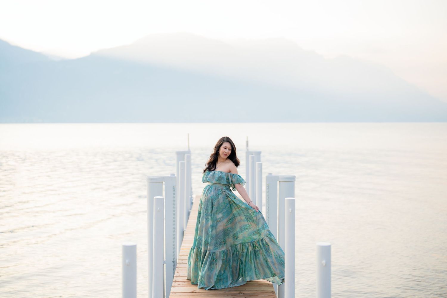 Woman in teal floral gown poses on white pier overlooking calm lake with misty mountains in background.