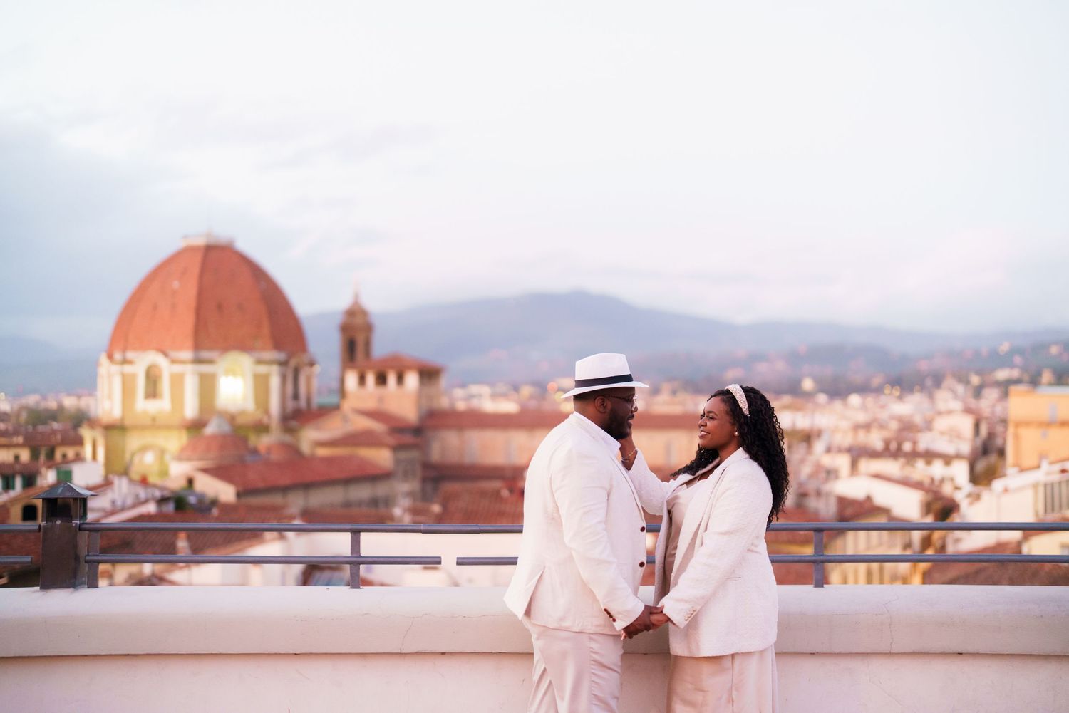 Couple in white attire holding hands on a rooftop in Florence, Italy, with the Duomo dome and cityscape behind them.