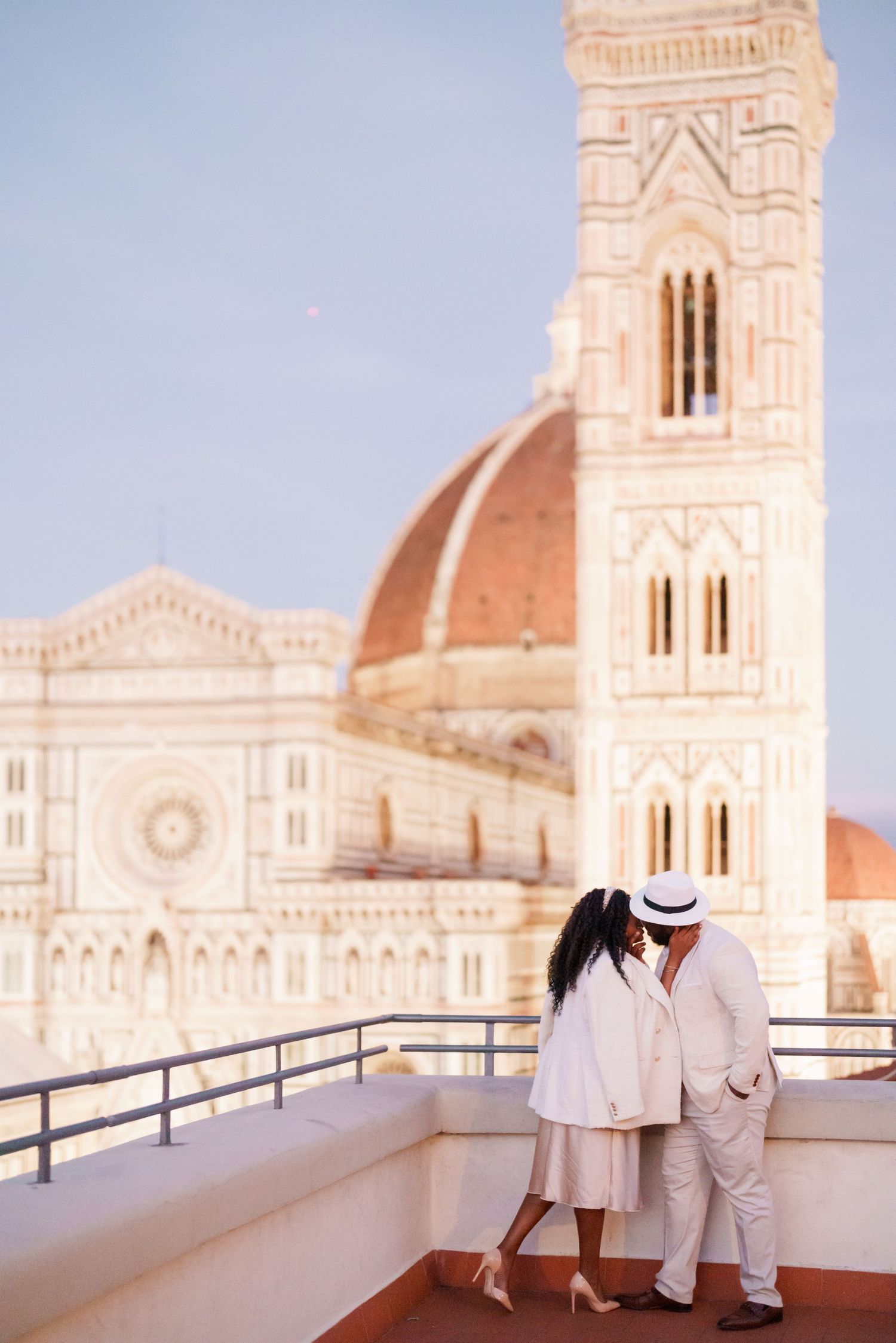 Romantic couple in white outfits kissing on a rooftop terrace with Florence Cathedral dome backdrop.