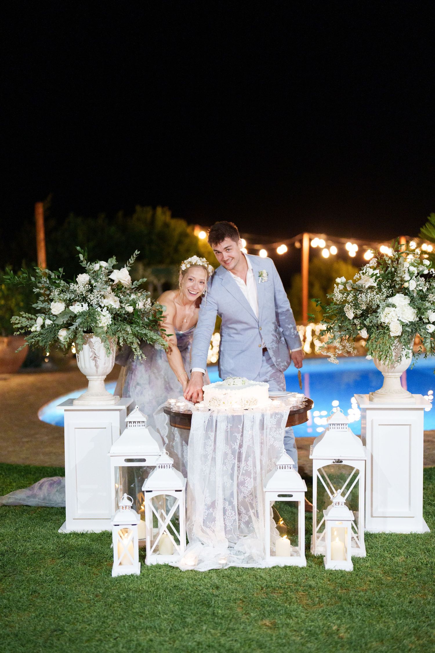 Bride and groom cutting wedding cake at night, surrounded by white lanterns and floral arrangements near a pool.