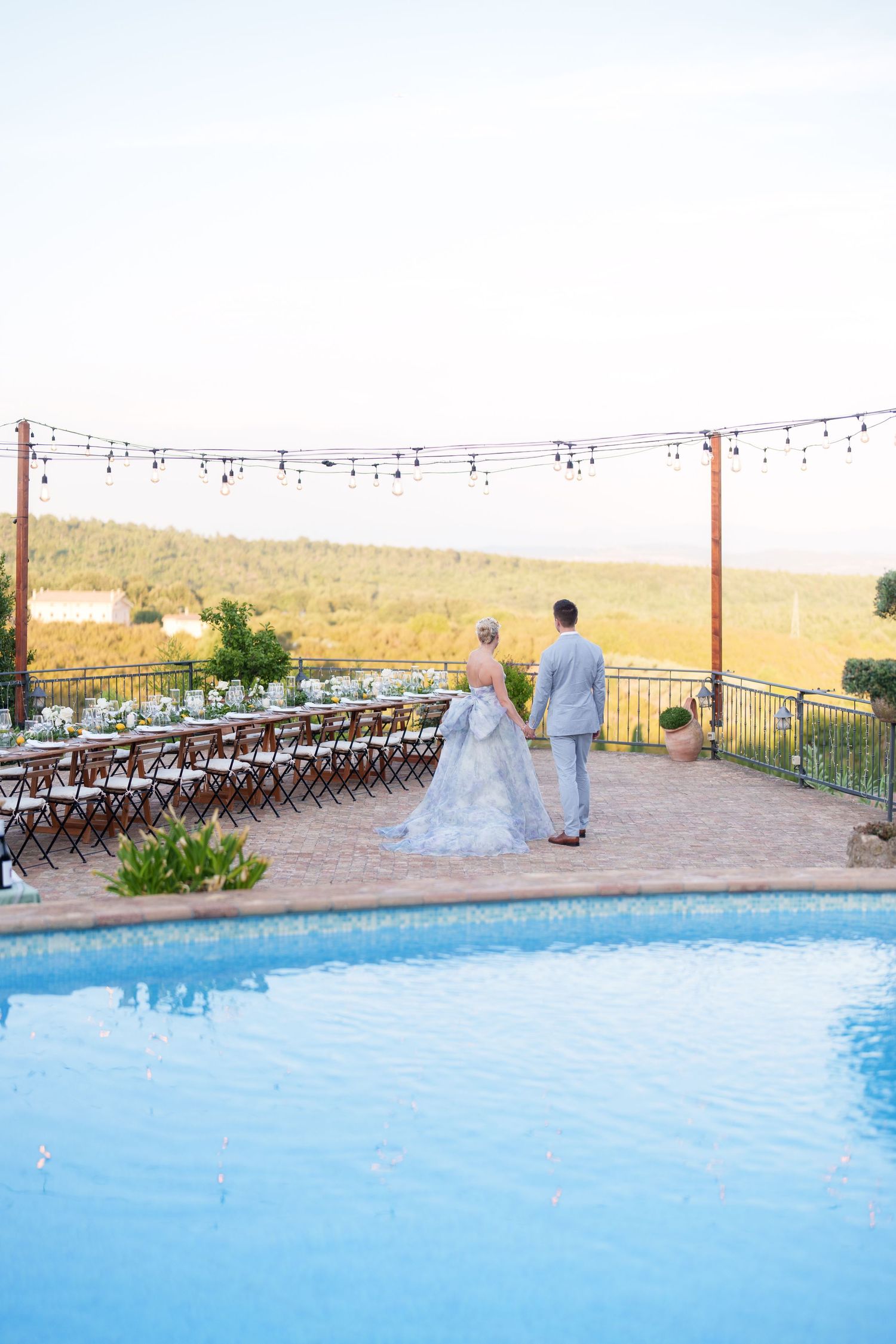 Bride and groom stand poolside at outdoor wedding reception with long dining table and string lights overlooking scenic vineyard.