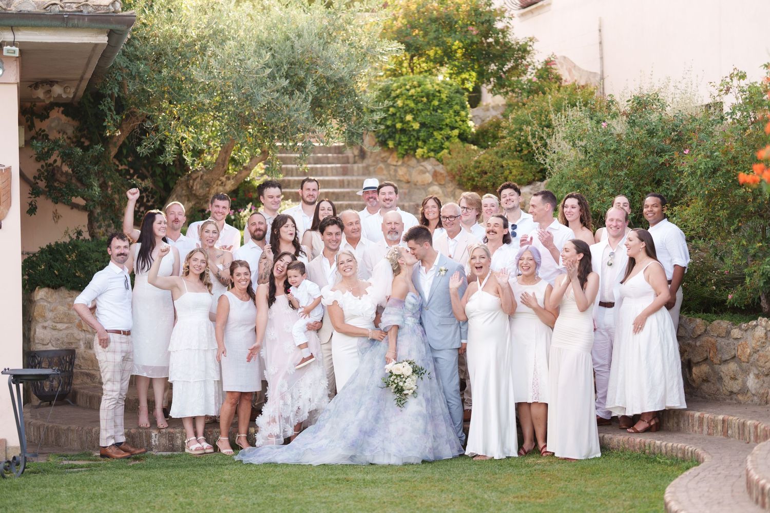 Large wedding party in white attire posing outdoors on a sunny day with lush greenery and stone steps in the background.