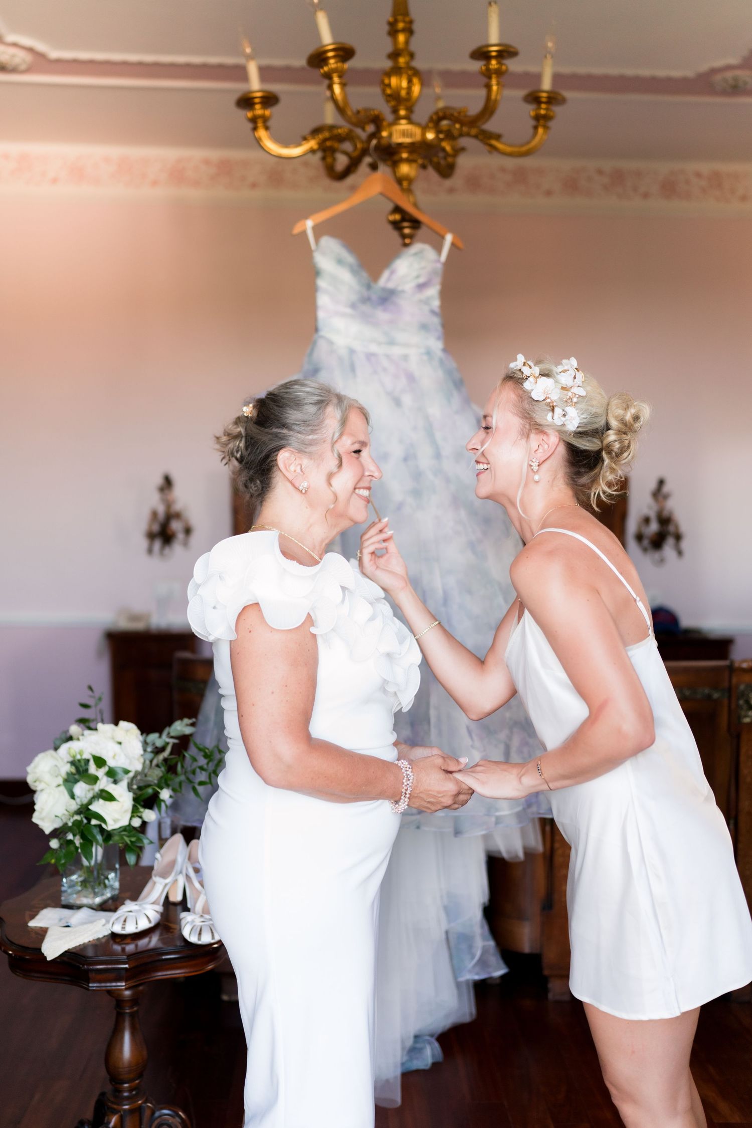Two brides in white dresses laugh together in an elegant room with a floral wedding gown hanging in the background.
