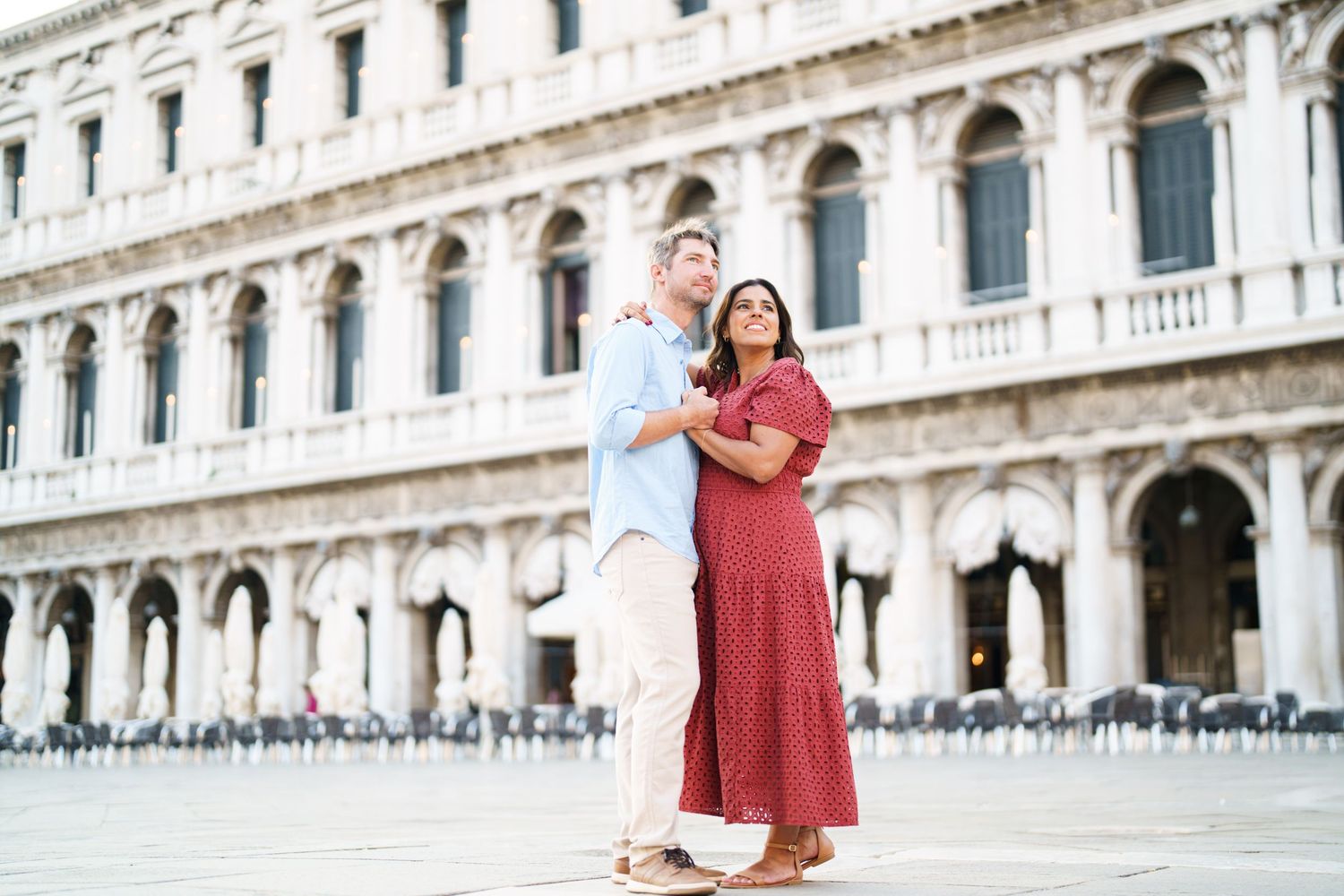 Couple embracing in St. Mark's Square, Venice, woman in red dress, man in light blue shirt, historic architecture behind.
