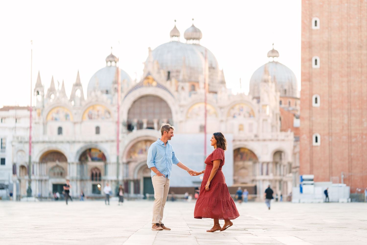 Couple holding hands in St. Mark's Square, Venice, with the ornate Basilica facade and campanile in the background.