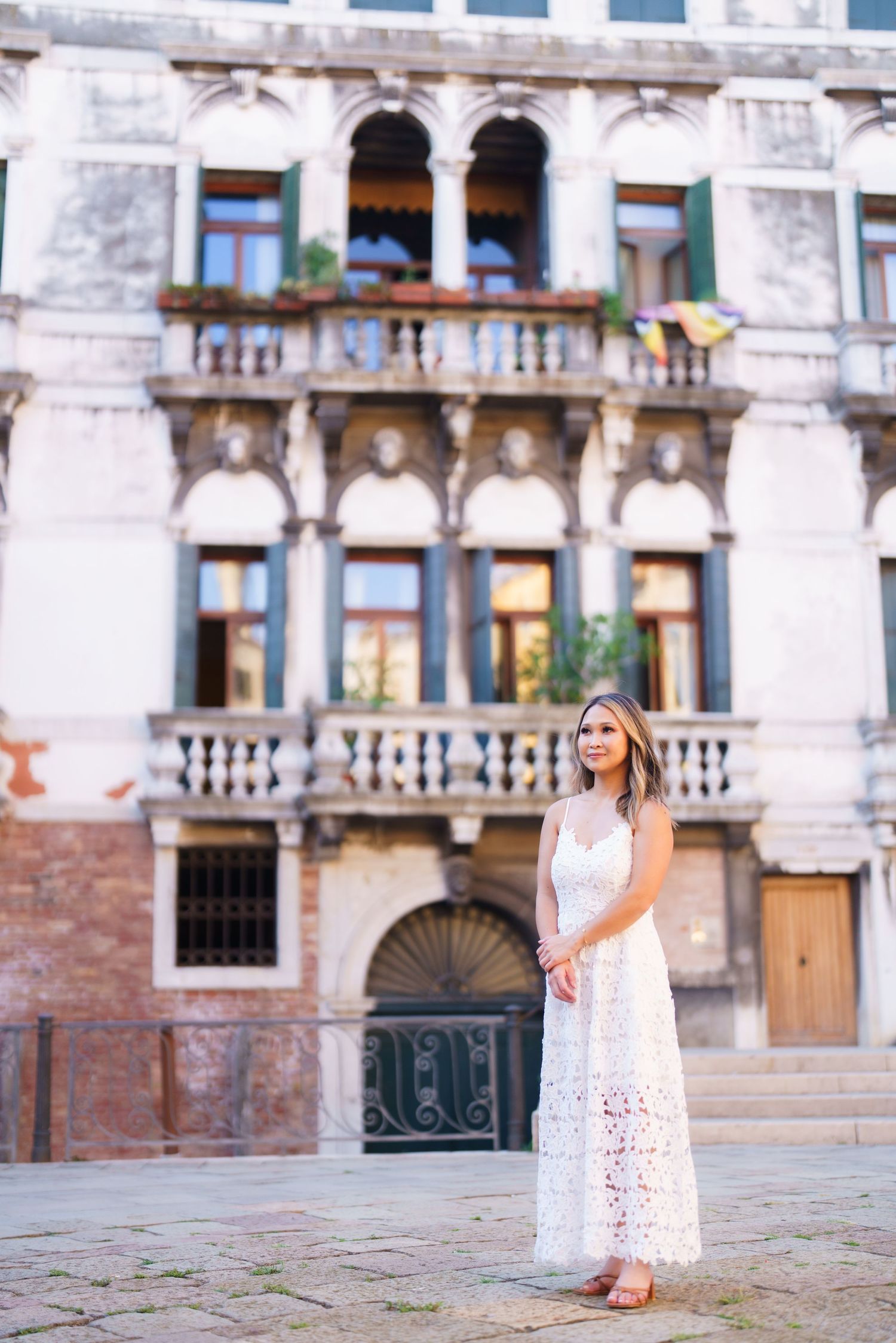 Woman in white lace dress standing before ornate Venetian palace with balconies in Italy.