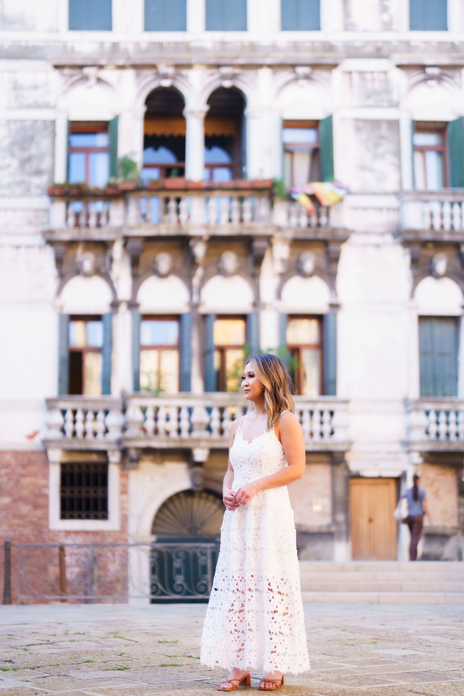 Woman in white lace dress standing in a Venetian piazza with ornate historic building facade behind her.