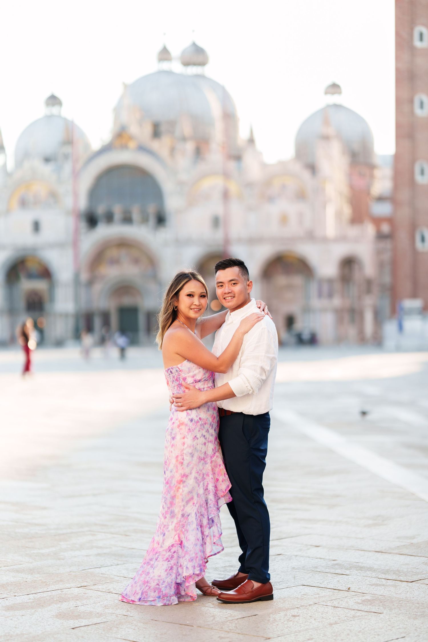 Couple embracing in floral dress and white shirt at St. Mark's Square, Venice, with the iconic basilica backdrop.
