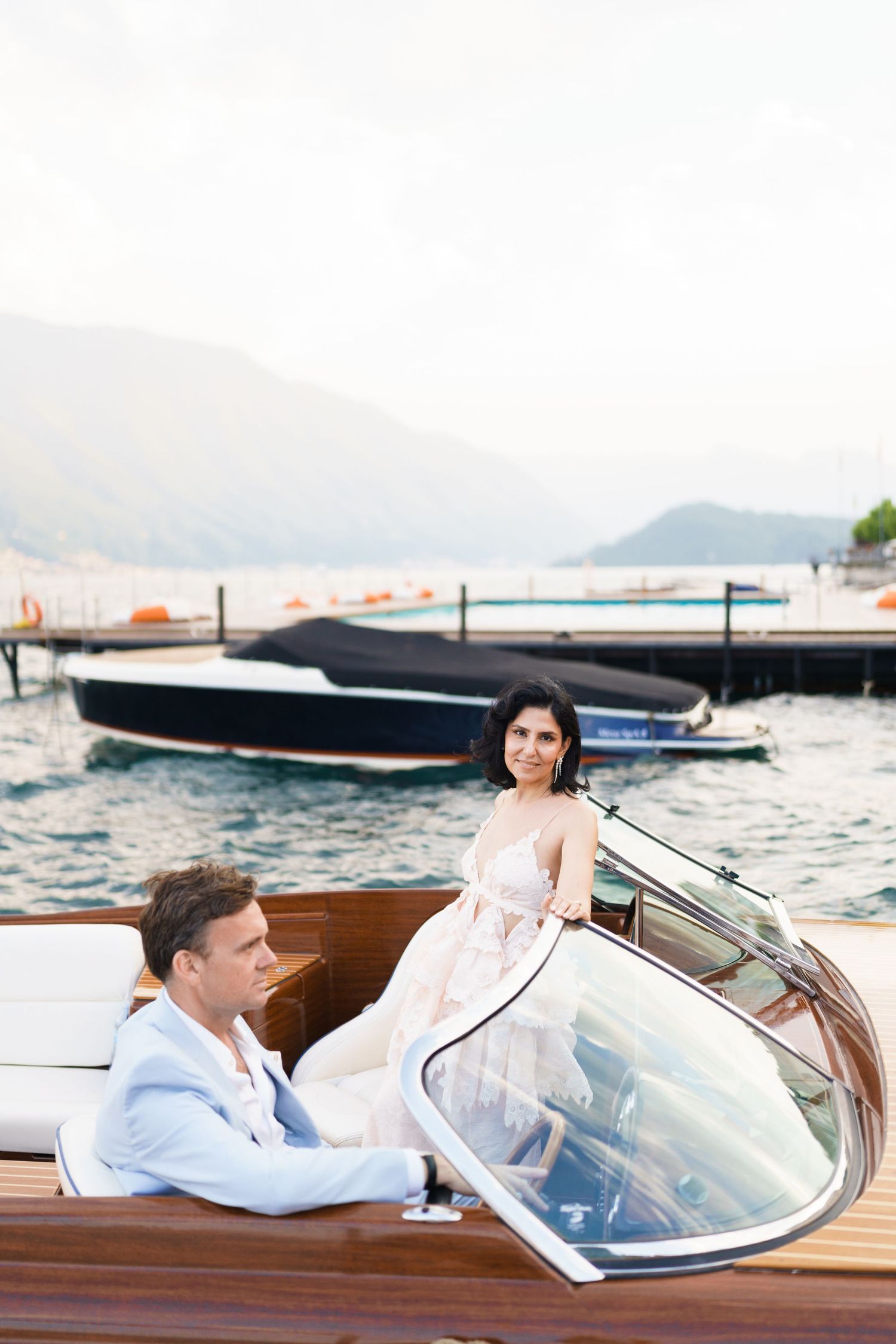 Elegant couple on a luxury wooden speedboat with mountain lake backdrop and marina in soft light.