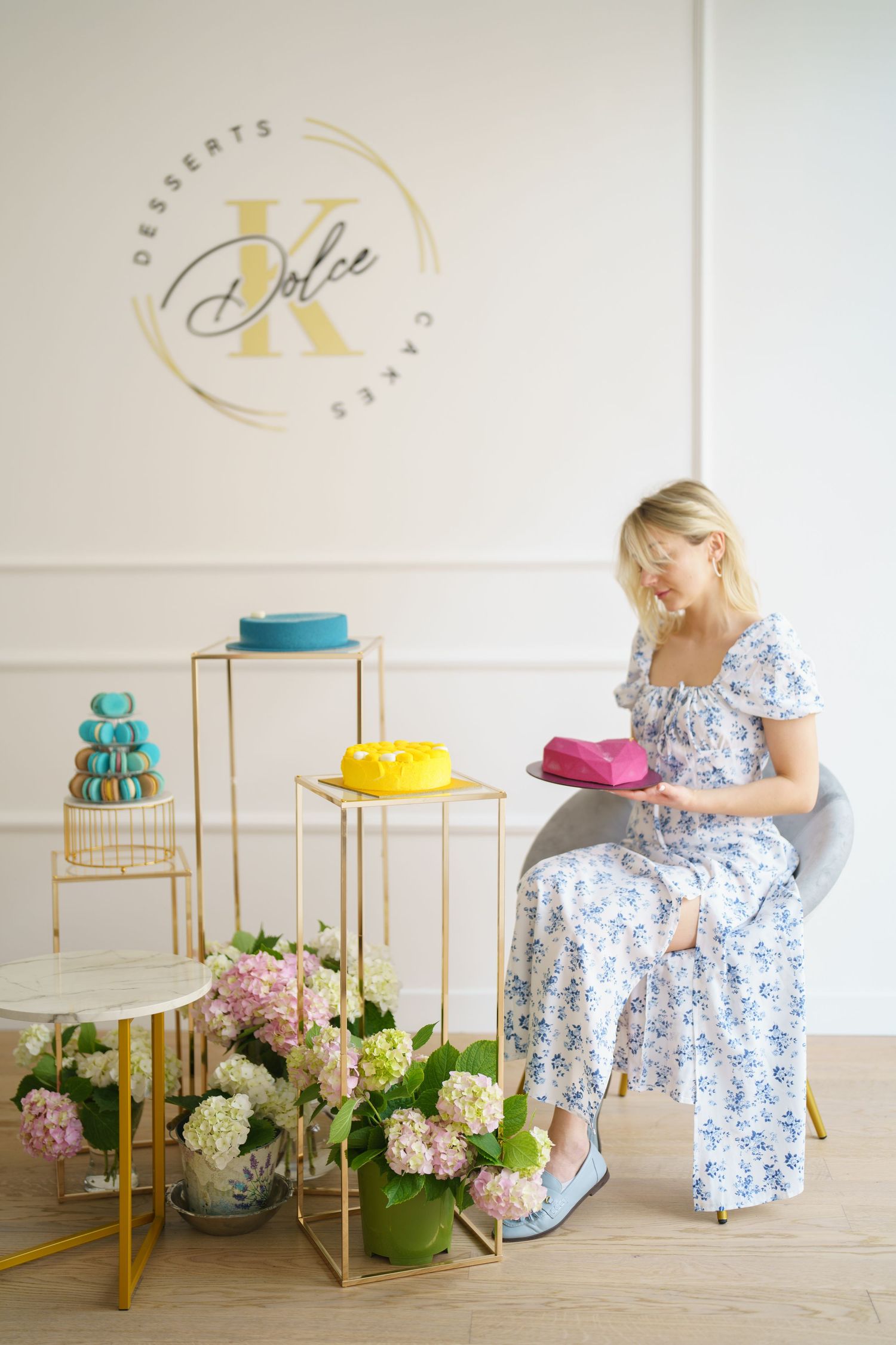 Woman in floral dress sits in bakery holding colorful cakes near hydrangeas, with Dolce K Desserts logo on wall.