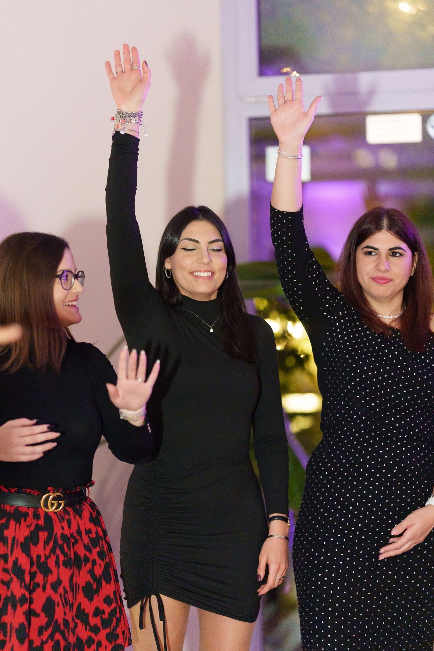 Three women dancing and waving hands at a party, wearing black and red outfits in an indoor venue.
