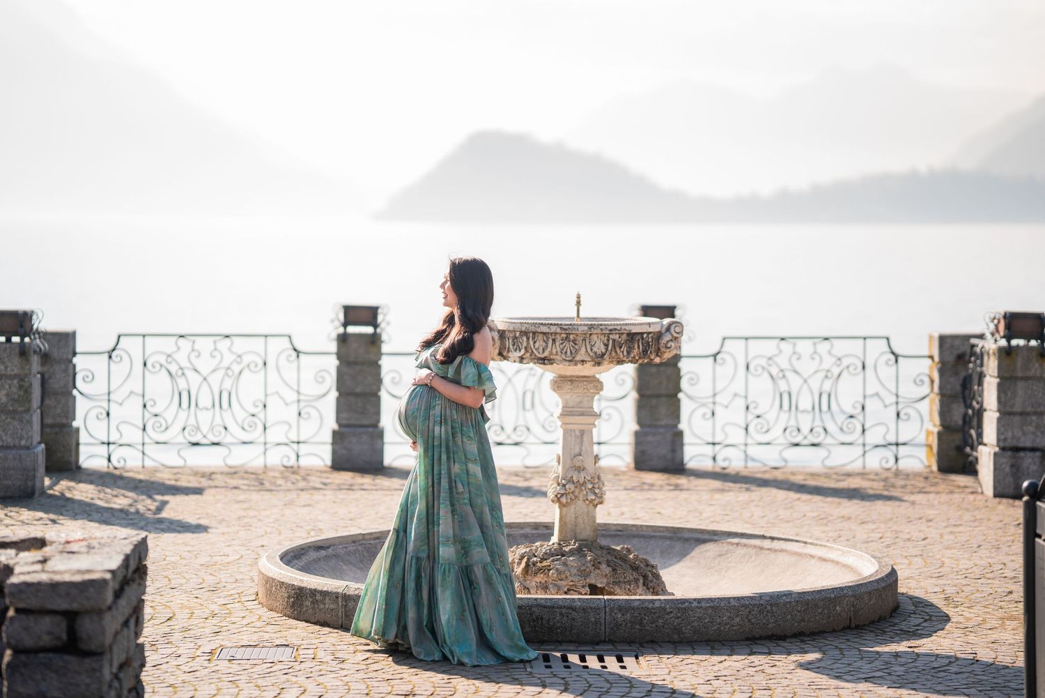 Pregnant woman in teal gown stands by stone fountain overlooking misty lake and mountains.