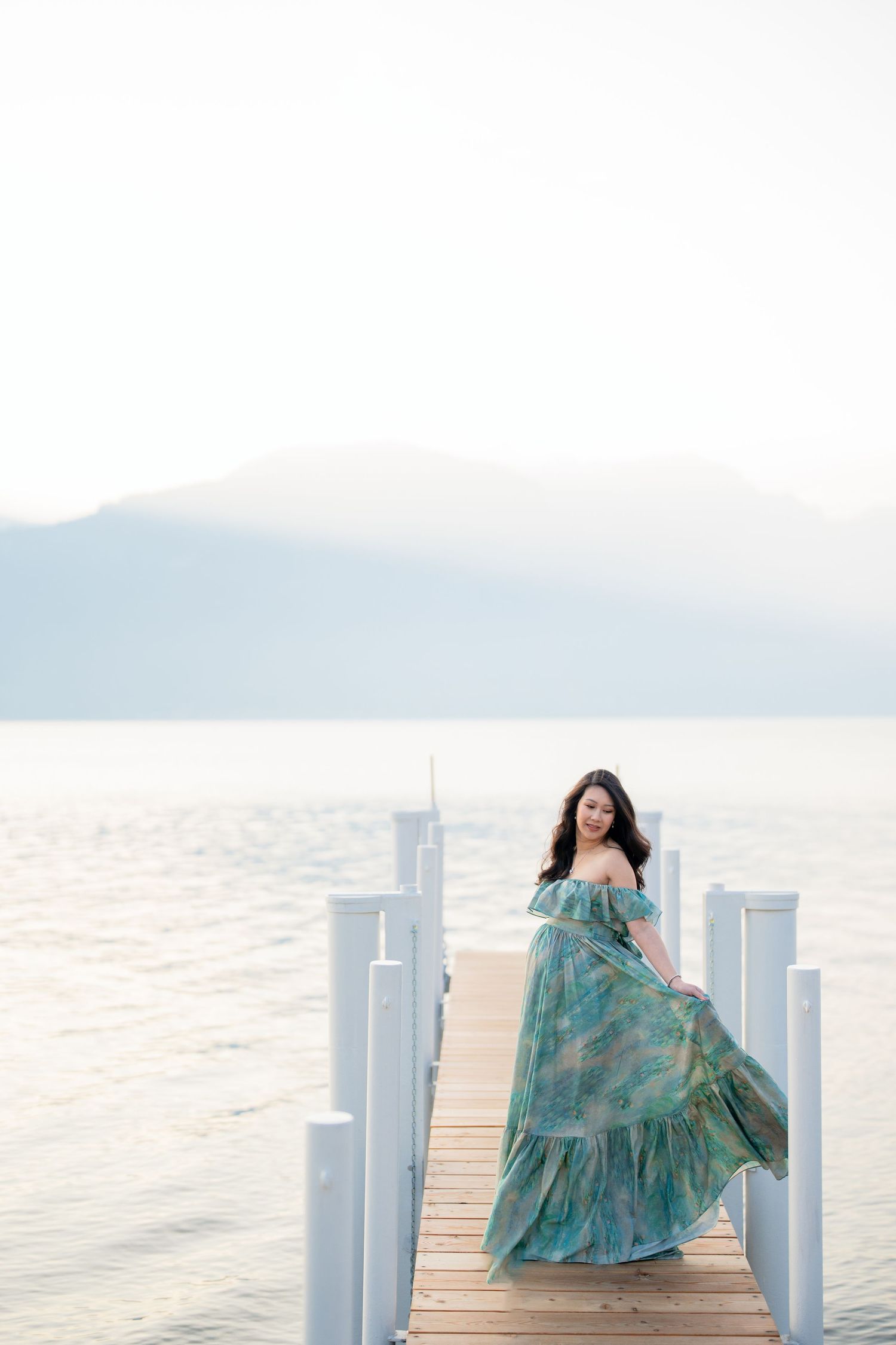 Woman in flowing teal floral gown poses on wooden dock with misty lake and mountains backdrop.