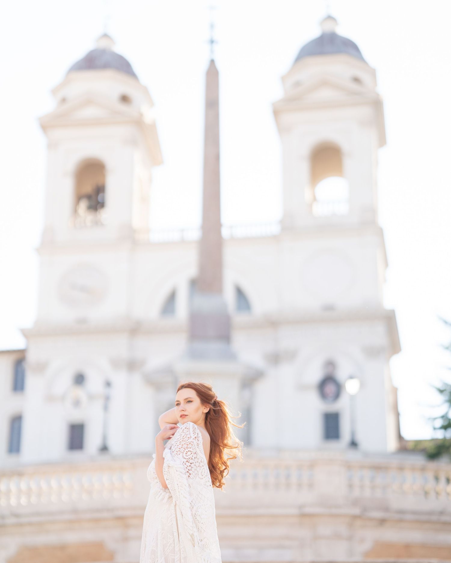 A woman in a white lace dress poses at the Spanish Steps in Rome with Trinità dei Monti church behind her.