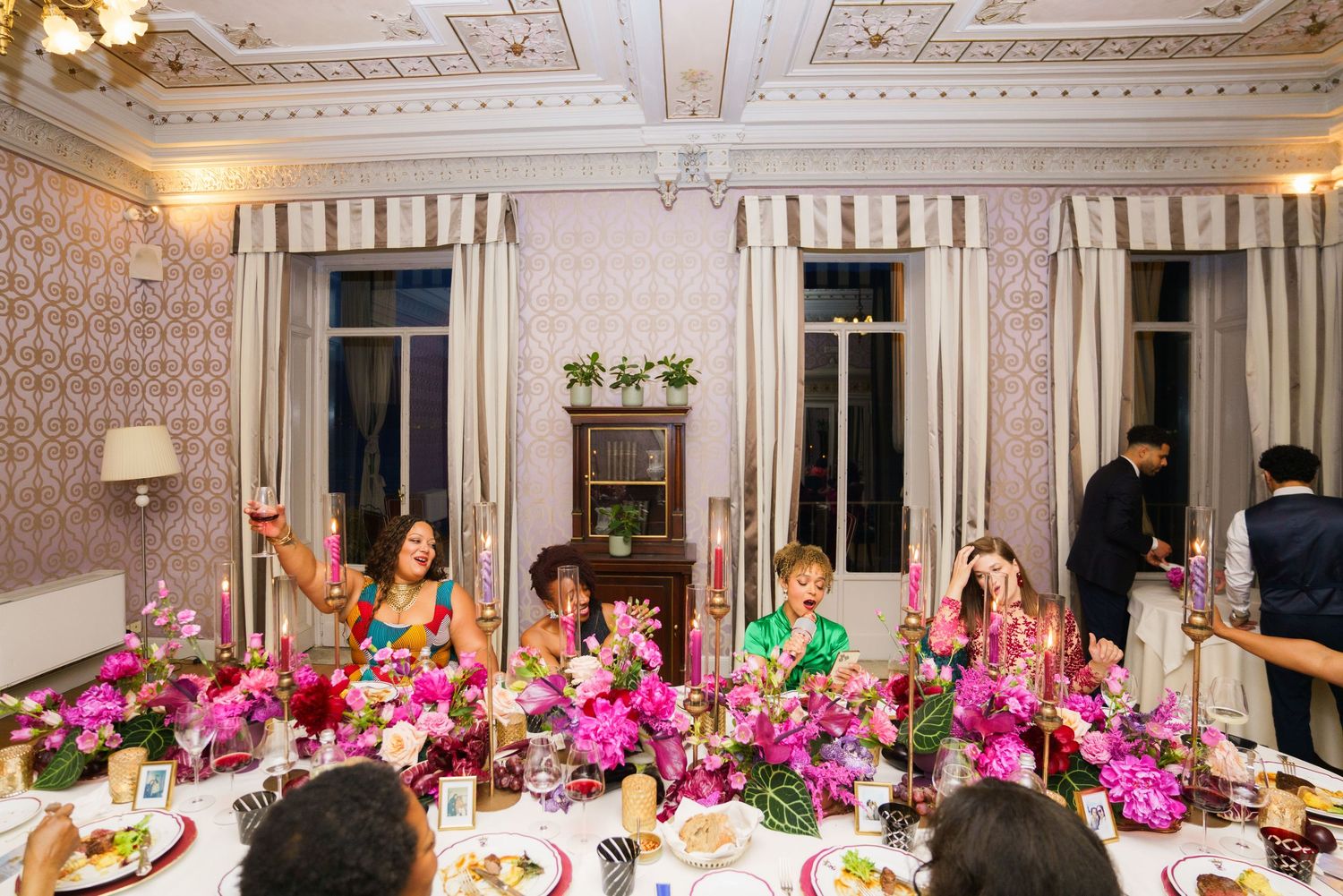 Elegant dinner party with guests seated at a floral-decorated table adorned with pink orchids and candles in a ornate room.