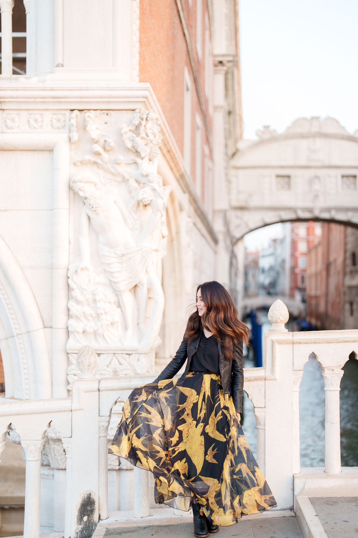 photographer venice Woman in flowing black and gold dress poses elegantly against ornate white marble columns in Venice.