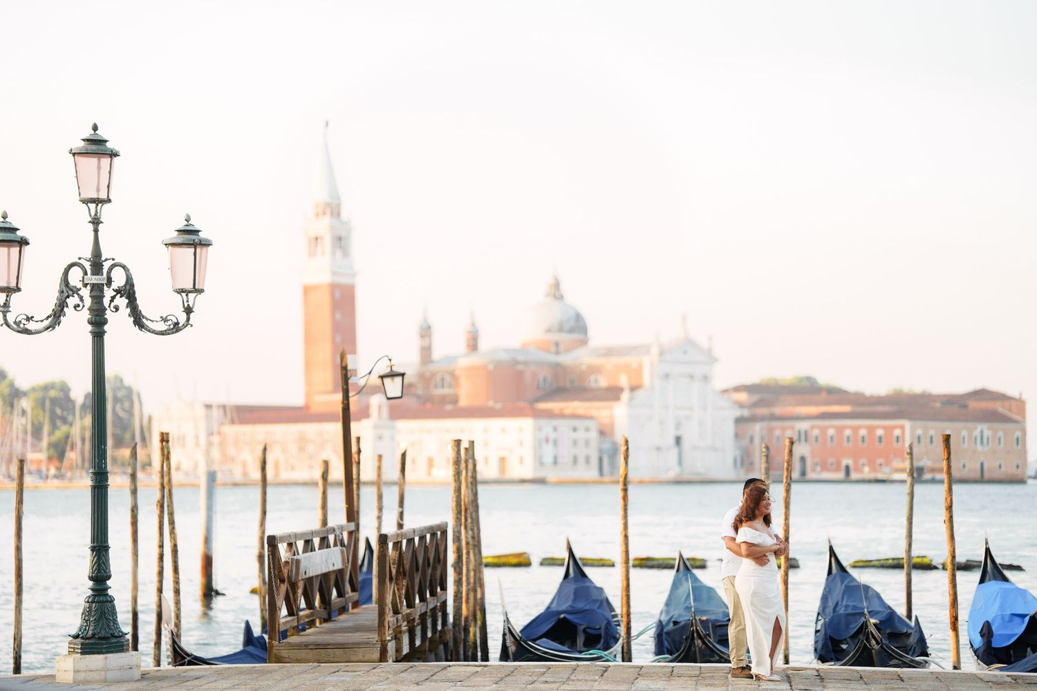 Panoramic view of Venice waterfront with covered gondolas and ornate lampposts along misty canal at dawn.