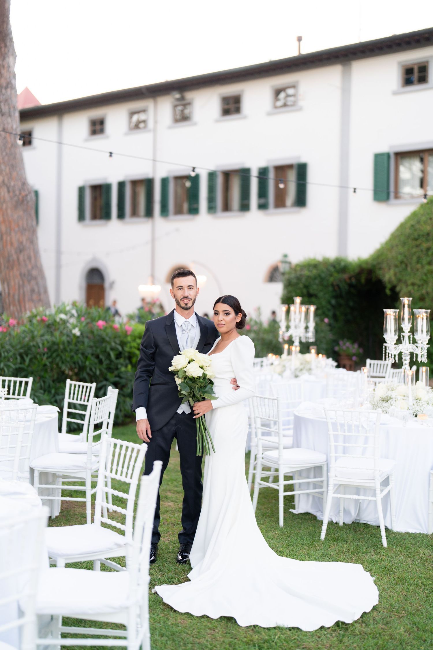 Elegant outdoor wedding reception setup with white chiavari chairs and tables arranged on a manicured lawn.