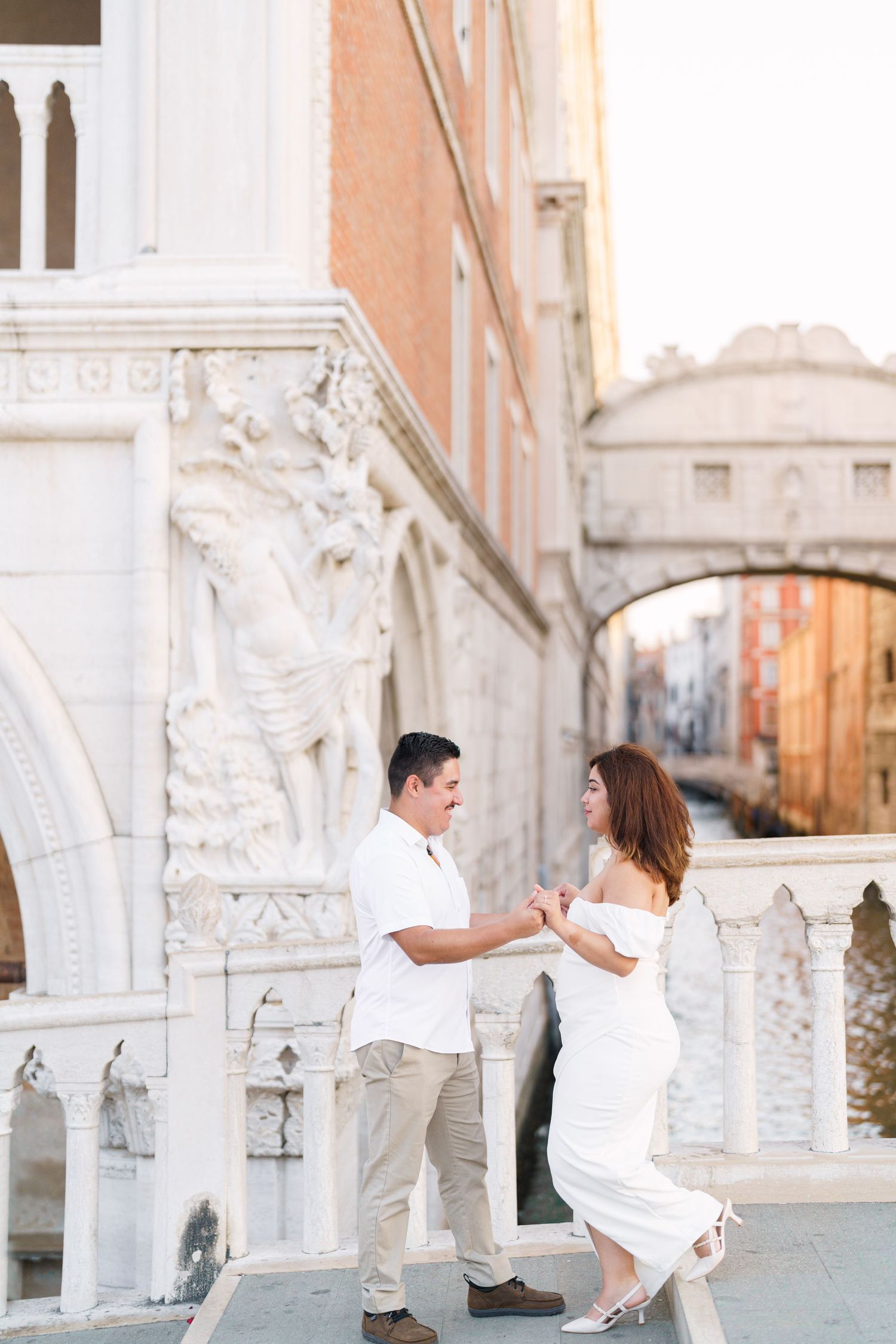 A couple in white outfits dances and twirls together in Venice's historic streets with ornate architecture in the background.