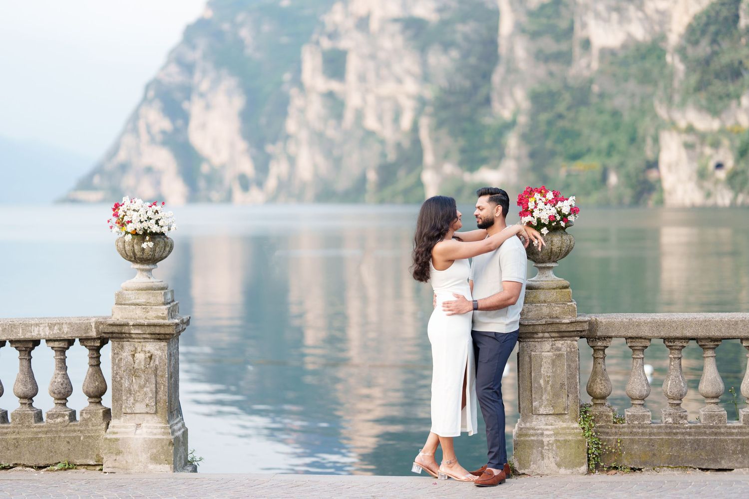 Romantic couple embraces on ornate stone balcony overlooking serene lake with mountain cliffs in background.