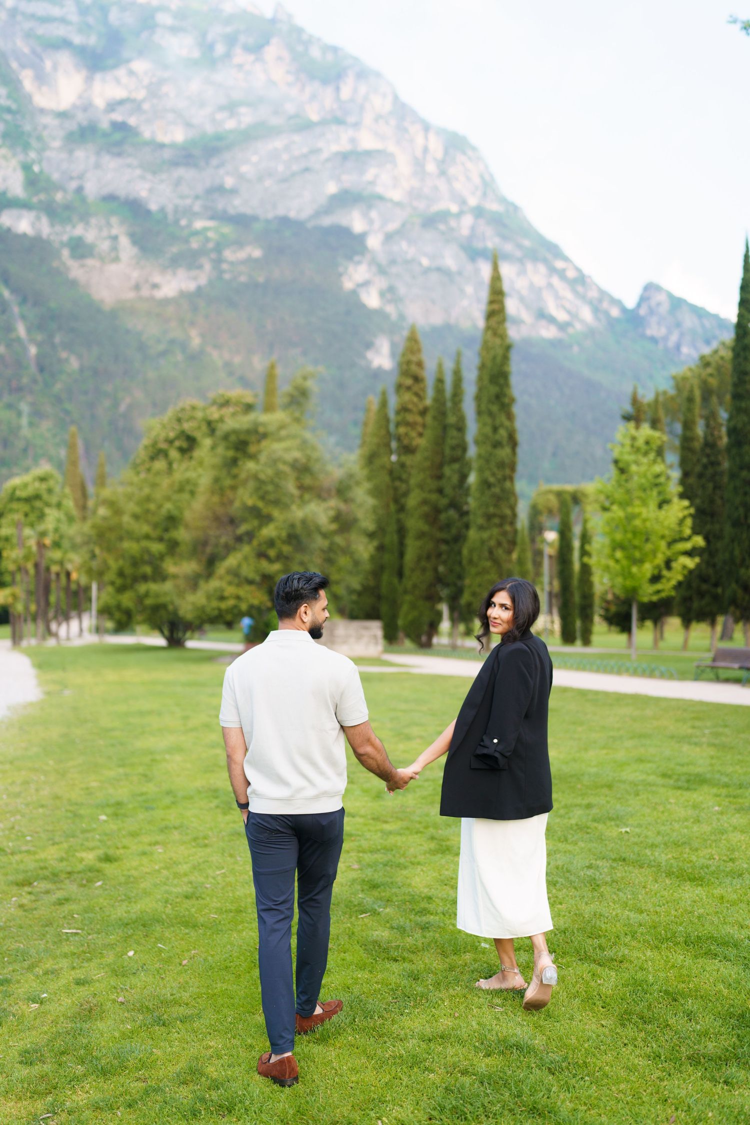 A couple walks hand in hand across a grassy meadow with majestic mountain peaks rising in the background.
