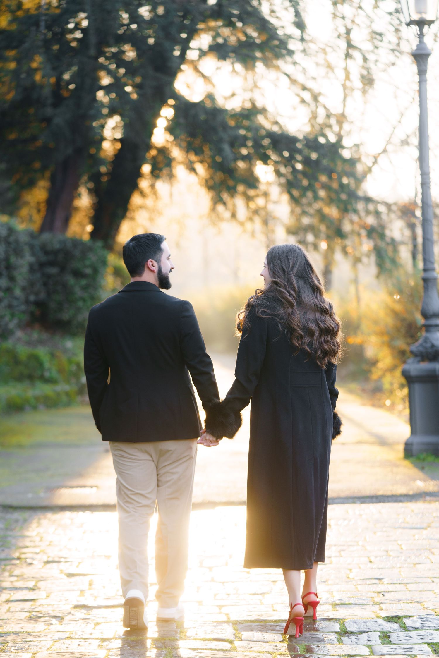 A couple walks hand in hand along a cobblestone path during golden hour with sunlight filtering through autumn trees.
