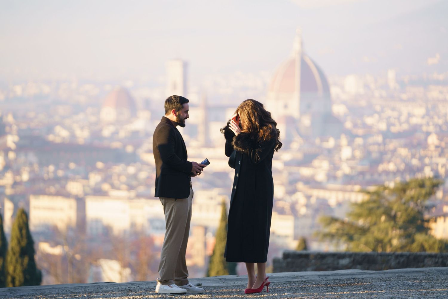 Two people stand on a hilltop overlooking Florence, Italy with the iconic Duomo cathedral visible in the misty cityscape.