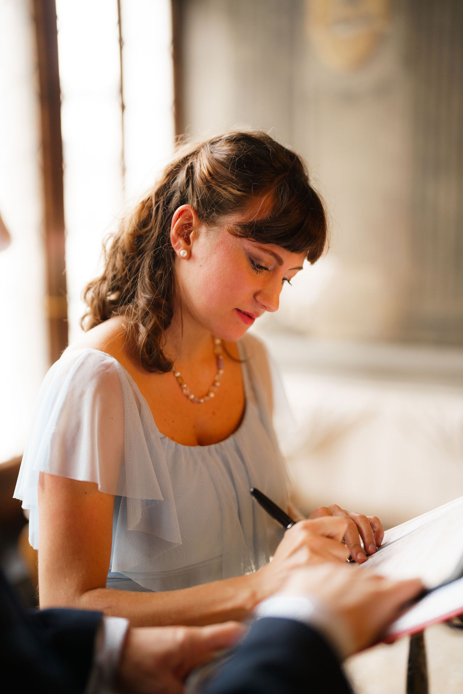 Someone in a white ruffled top signs a document while wearing a beaded necklace in warm natural lighting.
