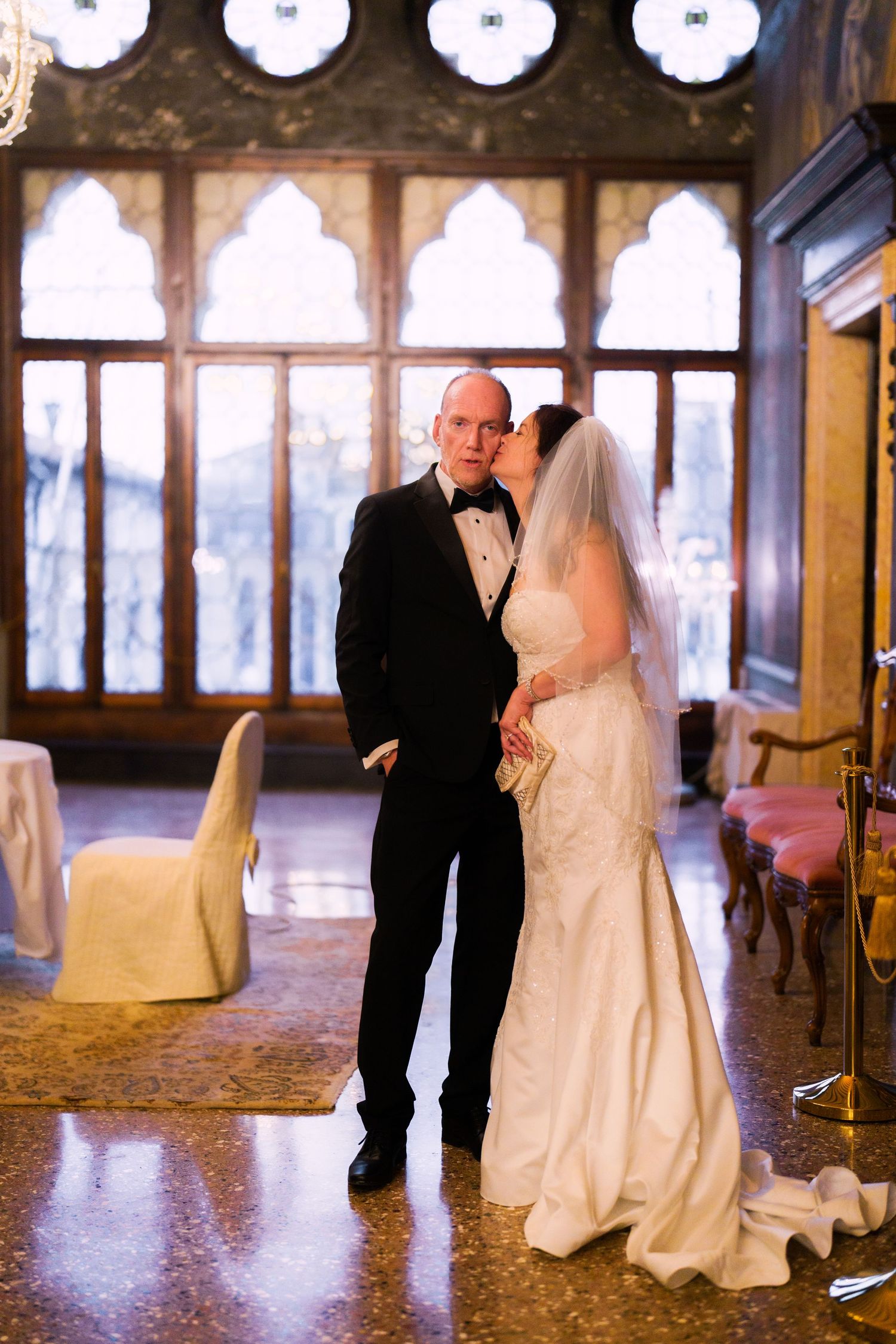 A bride and groom share a romantic dance in an elegant ballroom with ornate windows and warm lighting.