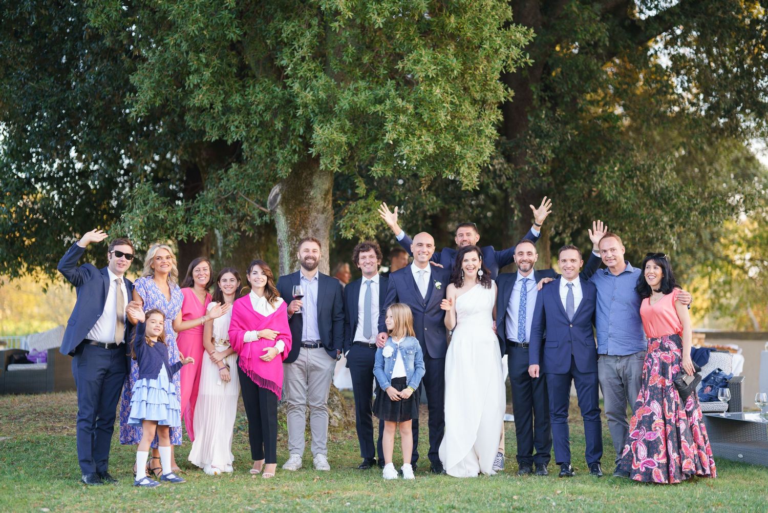 Large group photo of wedding guests and wedding party gathered outdoors under trees in formal attire.