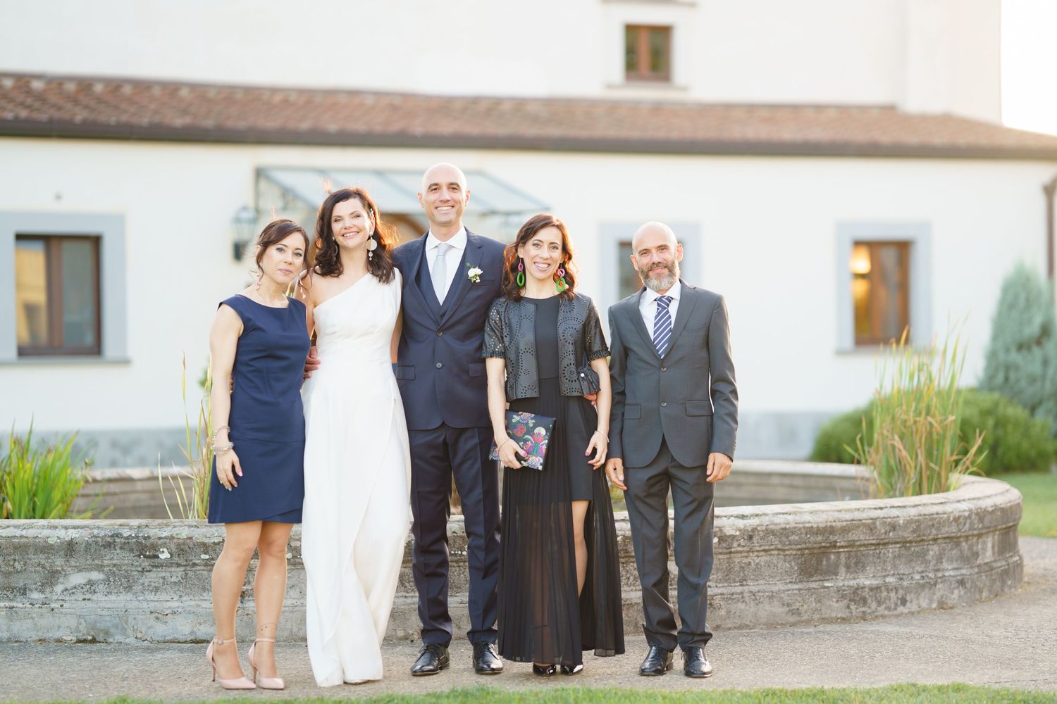 Wedding party members posing together outside white building with stone wall.