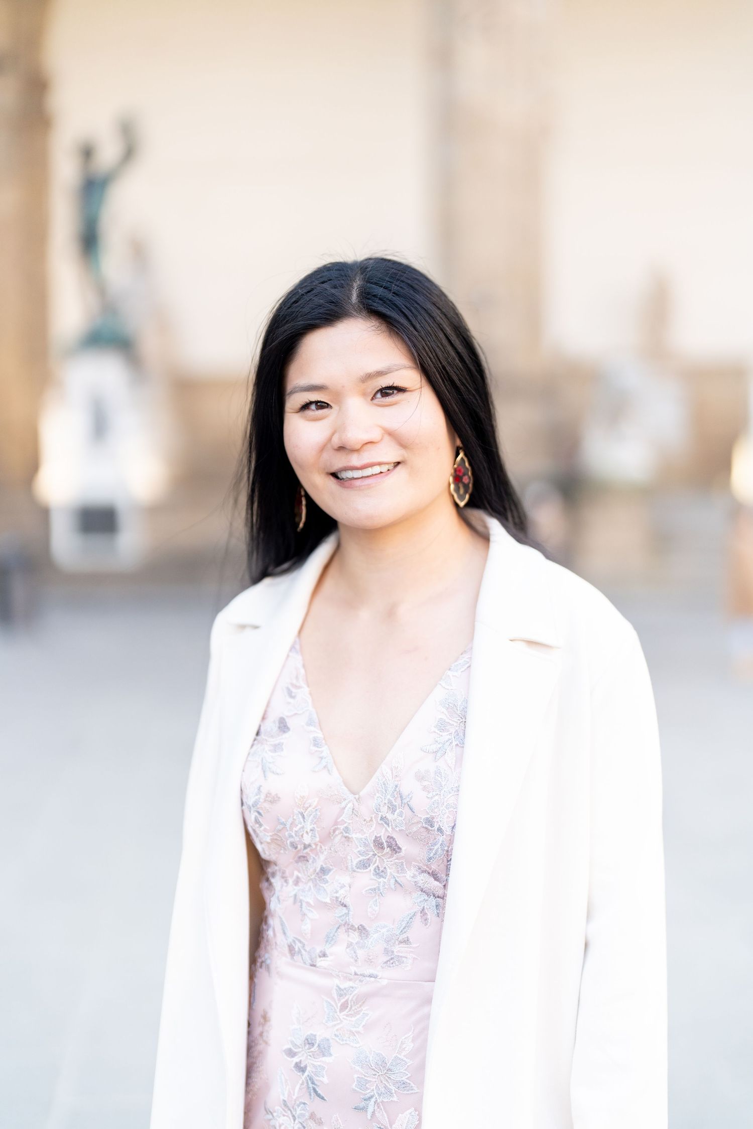 A person wearing a white blazer and floral dress smiles warmly while standing outdoors with a statue visible in background.