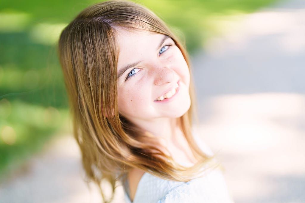 A person with long brown hair smiles joyfully in a sunny outdoor setting with green foliage in the background.