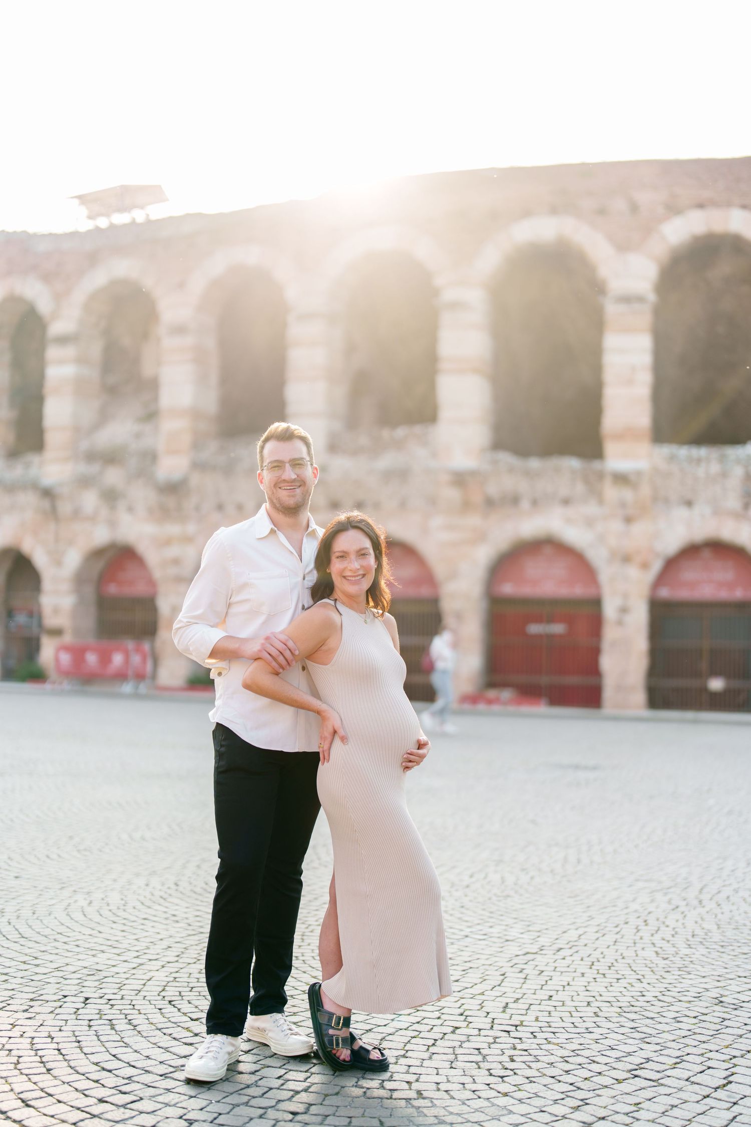 Couple in formal attire embraces on cobblestone plaza at sunset with arched architecture in background.
