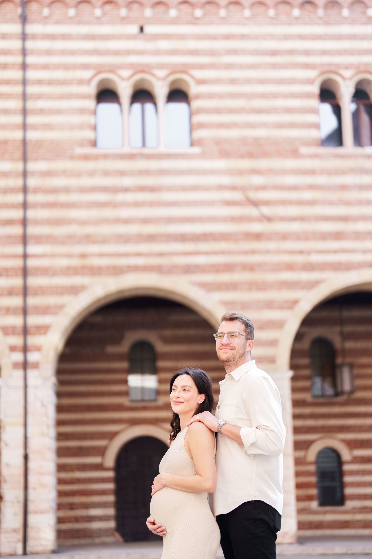 A couple stands together against a pink striped building with arched windows in a romantic architectural setting.