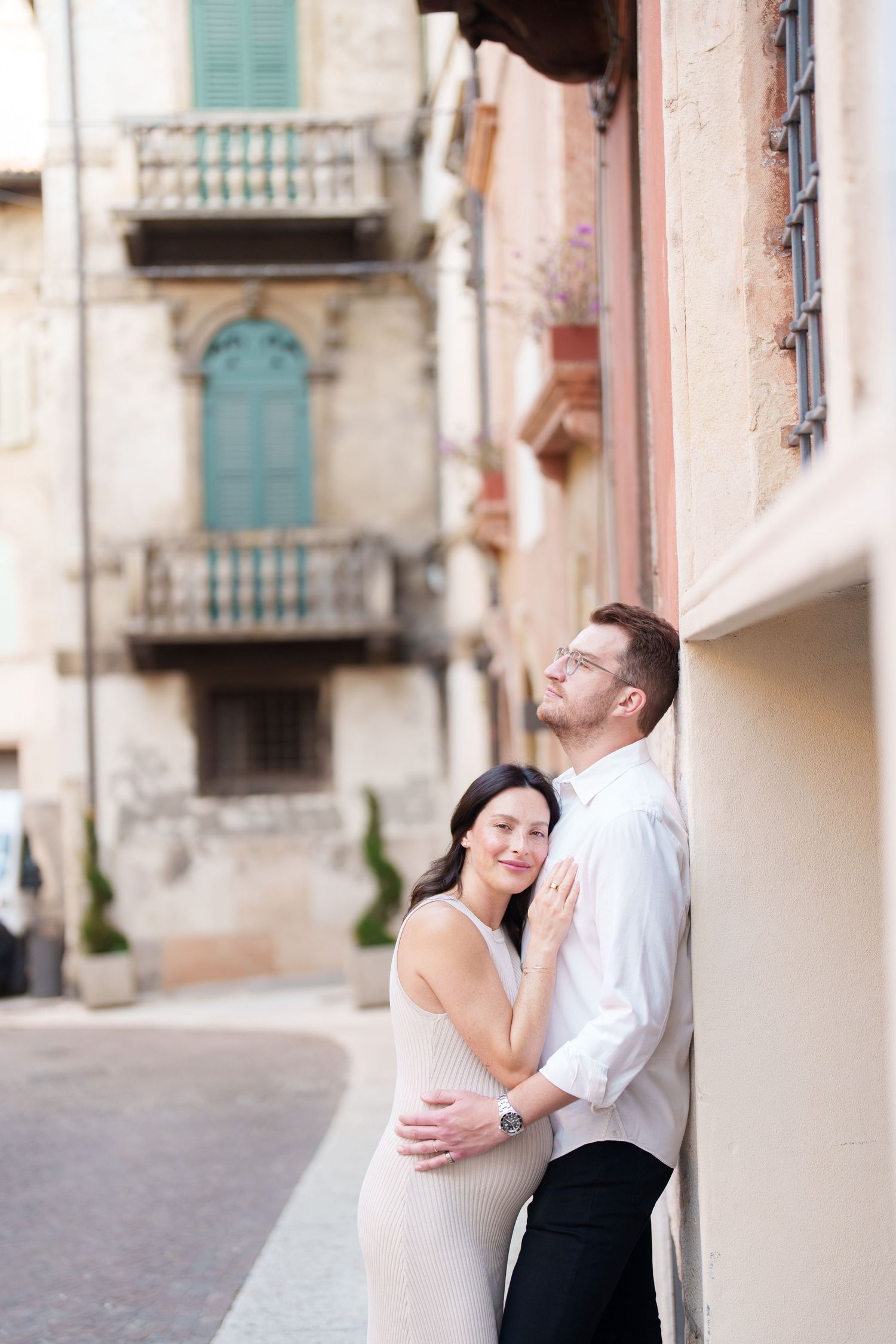A romantic couple embraces against a rustic European building facade with turquoise shutters and stone walls.