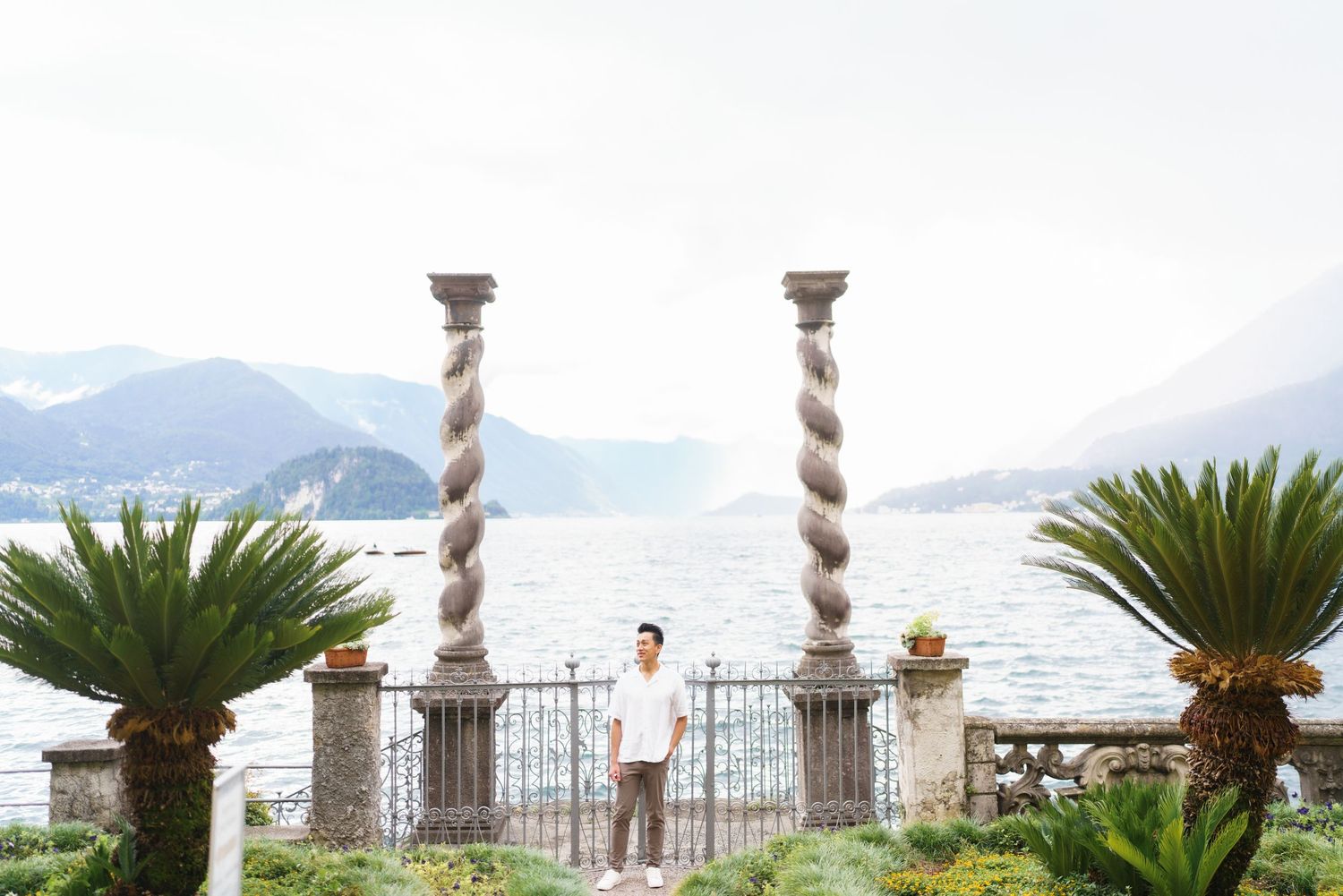 Ornate stone columns and palm trees line a Mediterranean waterfront terrace with mountains in the background.