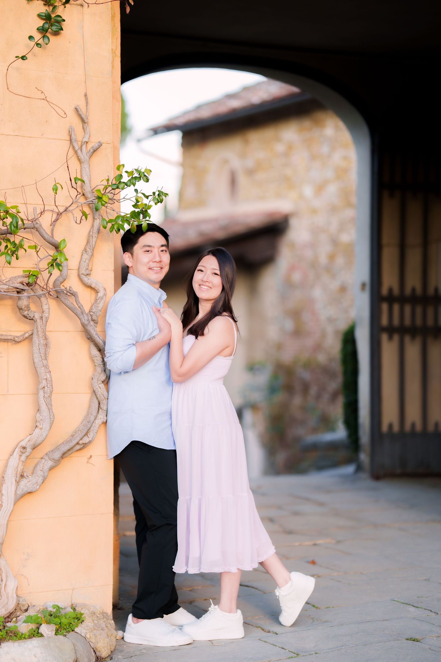A couple in light colored outfits poses together under an arched walkway with climbing vines at a rustic venue.