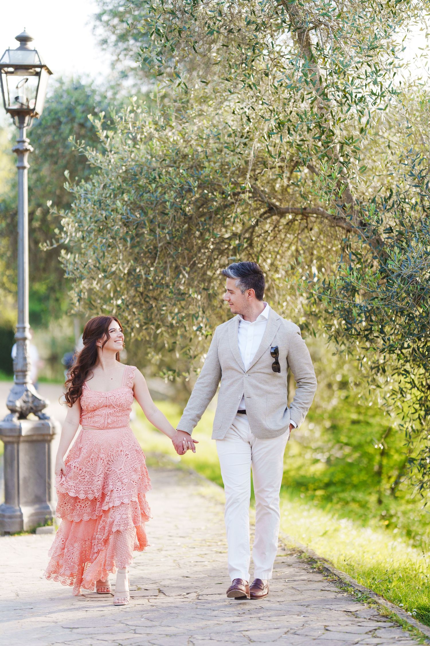 A couple in pink dress and white suit walk along a scenic path under olive trees and lamp posts.