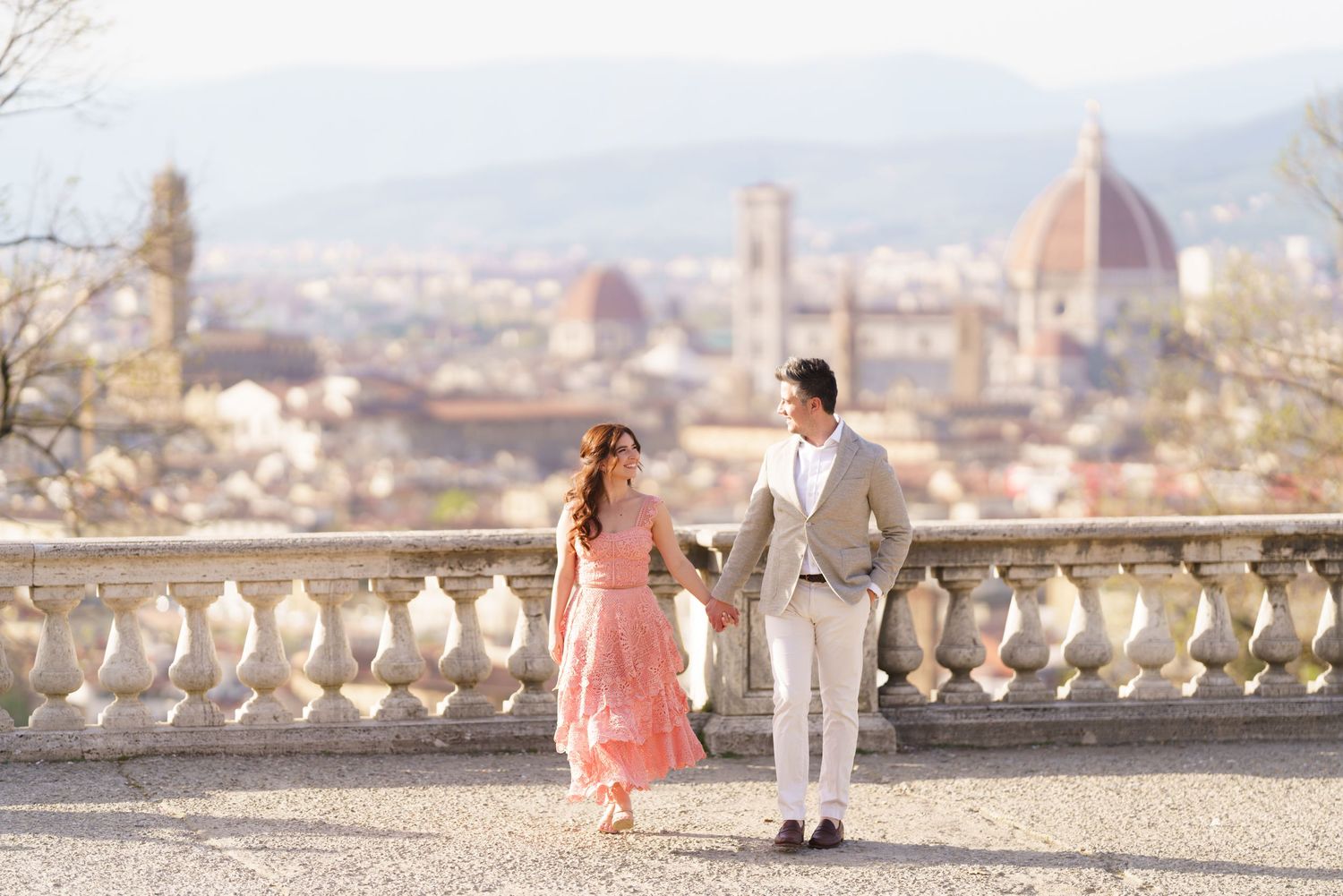 A couple in elegant attire walks along a scenic overlook with a view of Florence, Italy in the background.