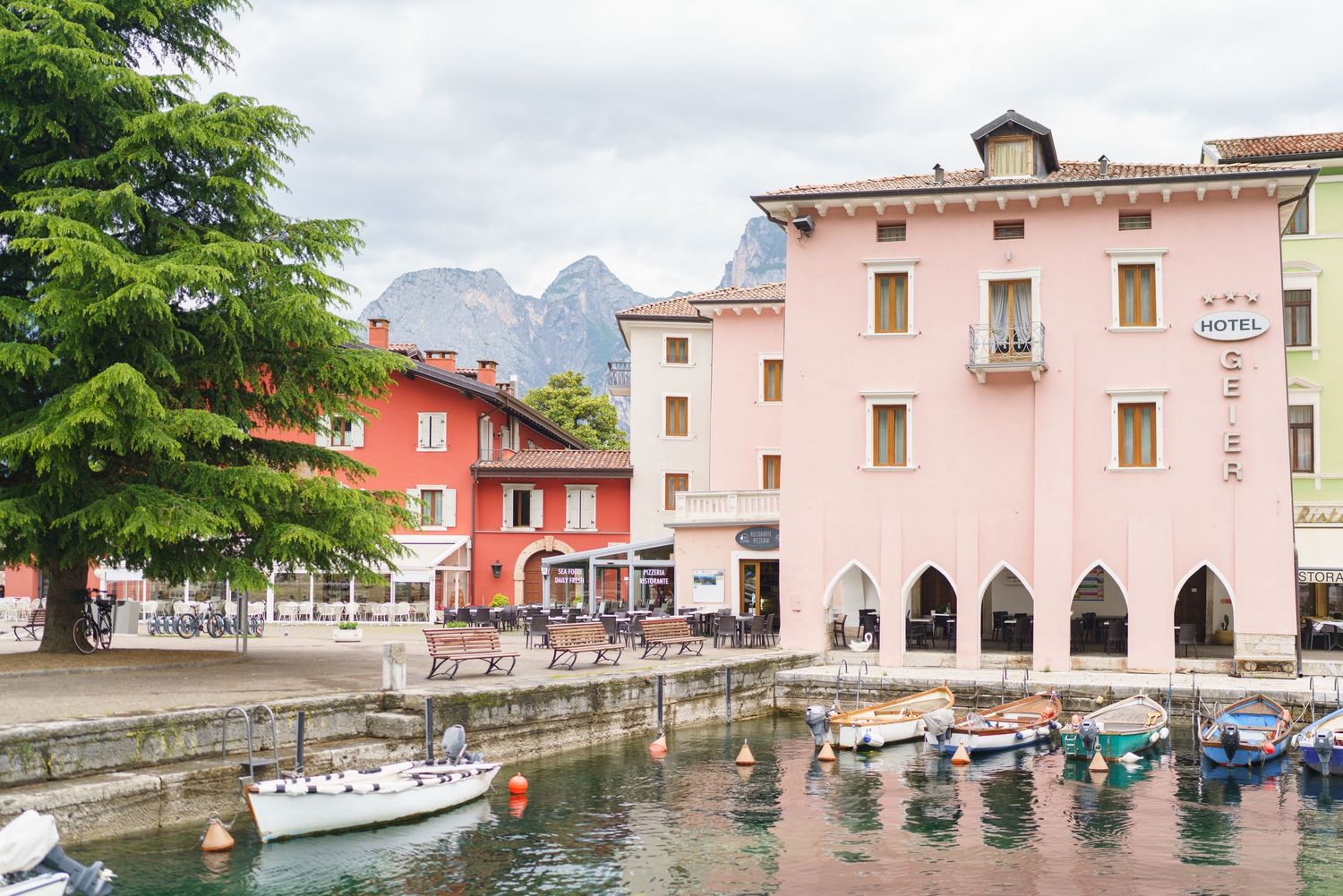 Scenic view of colorful buildings along a tranquil canal in Torbole, Lake Garda, Italy with boats and mountains in background.