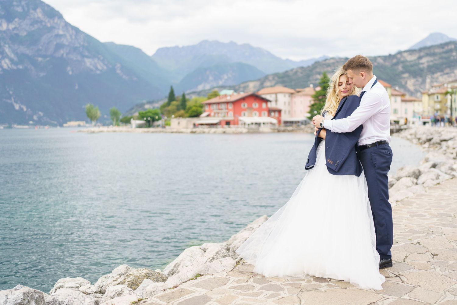 A couple shares a romantic moment on a scenic lakeside beach with mountains in the background.