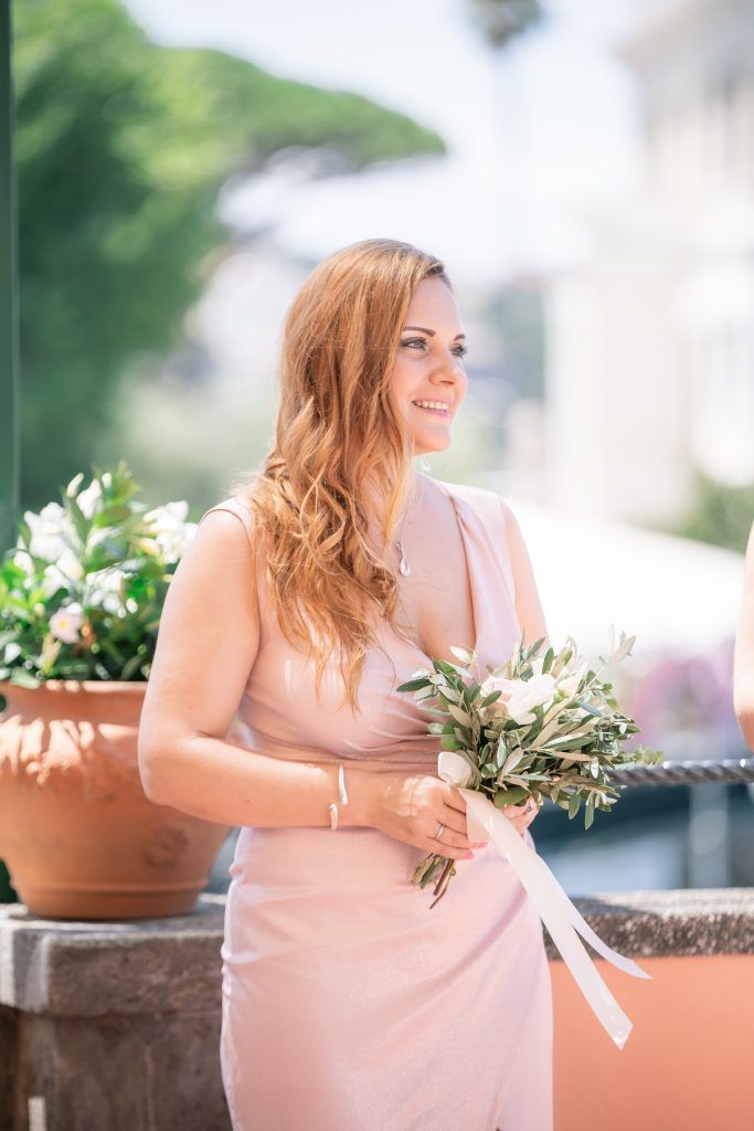 Bridesmaid in blush pink dress holds floral bouquet on sunny Mediterranean terrace.