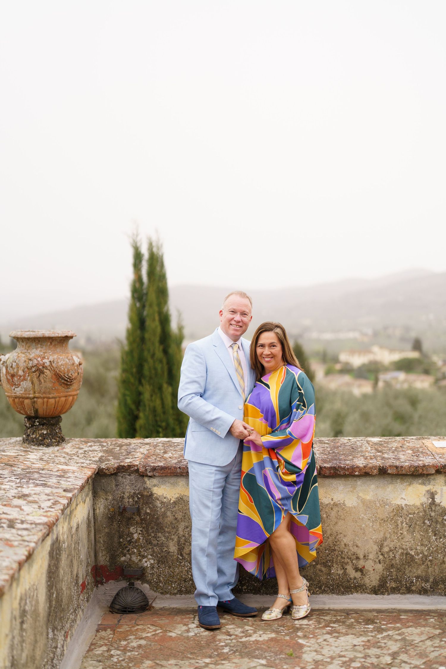 Couple in colorful attire sharing intimate moment on stone terrace overlooking Tuscan landscape.