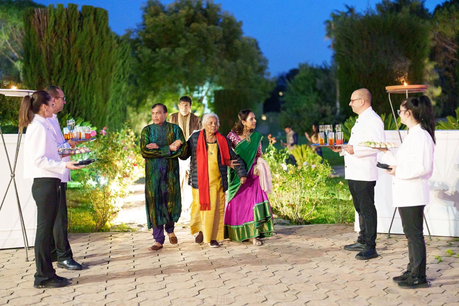 Evening garden party with guests walking along illuminated stone pathways.