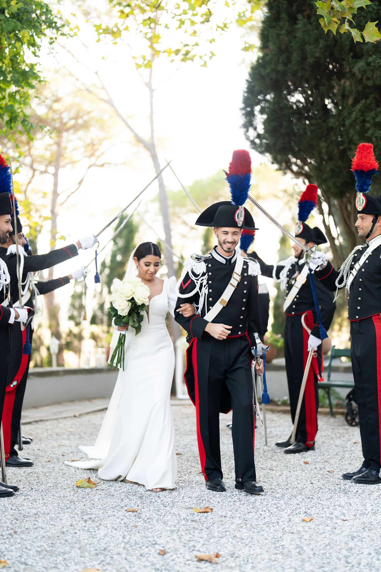 Military honor guard forms ceremonial line during outdoor wedding on gravel path.